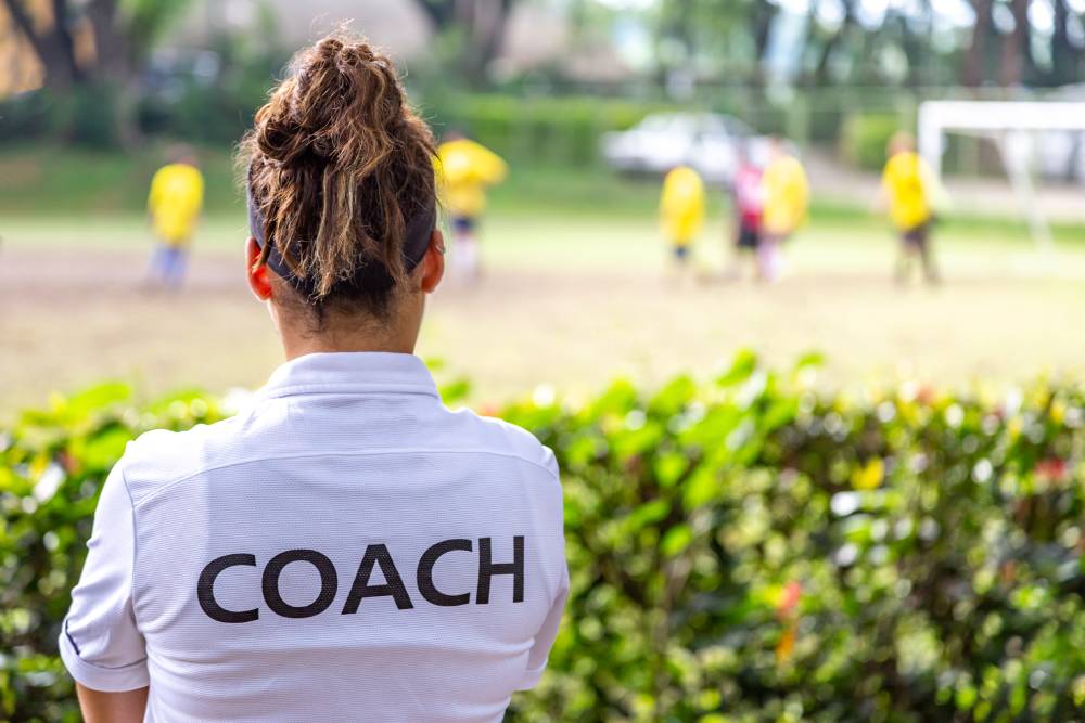 A woman's soccer coach watches her team from the sideline.