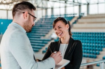 Sports marketing professionals stand in a stadium and laugh in enjoyment as they stand in an arena planning an event.